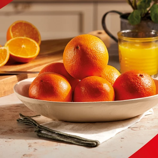 Laranjas colocadas numa fruteira de cerâmica com um copo de sumo de laranja ao fundo